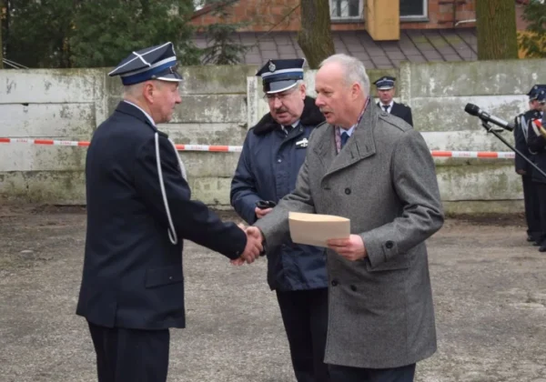 Uroczystość przekazania samochodu dla OSP Mała Wieś: dwie osoby, jedna w mundurze, druga w eleganckim płaszczu, ściskają sobie dłonie i trzymają dokument, w tle stoją inni funkcjonariusze.
