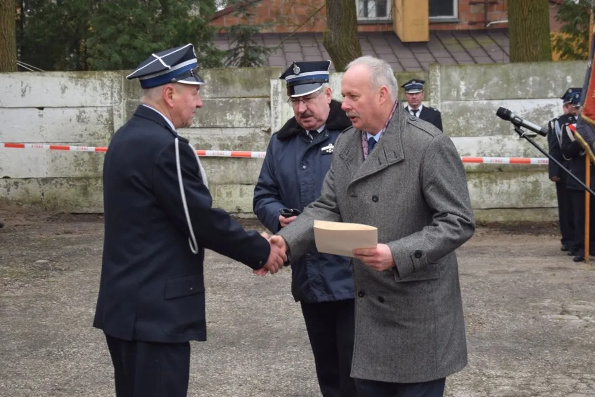 Uroczystość przekazania samochodu dla OSP Mała Wieś: dwie osoby, jedna w mundurze, druga w eleganckim płaszczu, ściskają sobie dłonie i trzymają dokument, w tle stoją inni funkcjonariusze.