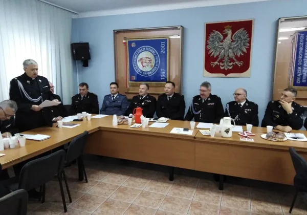 Członkowie zarządu Oddziału Powiatowego ZOSP RP w Płocku podczas plenarnie­go posiedzenia siedzą przy długim stole w sali konferencyjnej, a jeden z nich stoi i odczytuje dokument.