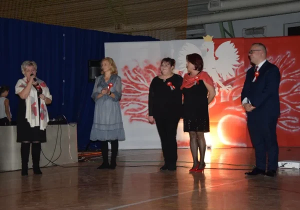 Pięć dorosłych osób stoi na scenie podczas szkolnej uroczystości z okazji 100-lecia odzyskania niepodległości w Szkole Podstawowej w Wyszogrodzie; prowadząca z mikrofonem po lewej, pozostali w eleganckich ubraniach z czerwonymi rozetkami, na tle czerwono-białego banneru z orłem.