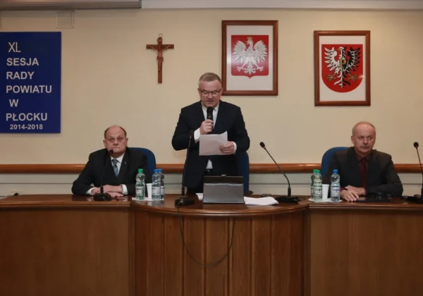 Trzy osoby na sali obrad Powiatu Płockiego podczas ostatniej sesji kadencji 2014–2018; mężczyzna stojący odczytuje z kartki, a dwóch siedzi przy stole, za nimi widoczne są krzyż, dwa herby i baner z napisem XL Sesja Rady Powiatu w Płocku 2014–2018.