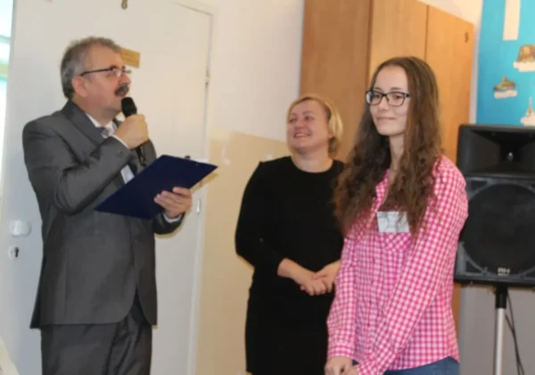 Mężczyzna w garniturze wygłasza przemówienie do mikrofonu podczas podsumowania projektu Stowarzyszenia „Edukacja dla Drobina”; obok niego stoją dwie kobiety, a młoda dziewczyna w koszuli w kratę.