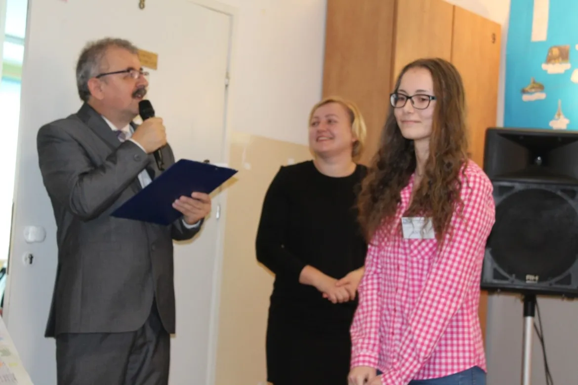 Mężczyzna w garniturze wygłasza przemówienie do mikrofonu podczas podsumowania projektu Stowarzyszenia „Edukacja dla Drobina”; obok niego stoją dwie kobiety, a młoda dziewczyna w koszuli w kratę.