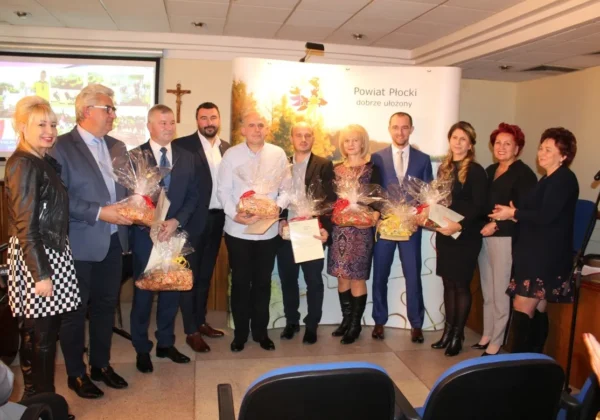 Grupa osób w eleganckich strojach stoi na sali podczas podsumowania współzawodnictwa sportowego szkół w powiecie płockim, trzymając kosze z upominkami i dokumenty.