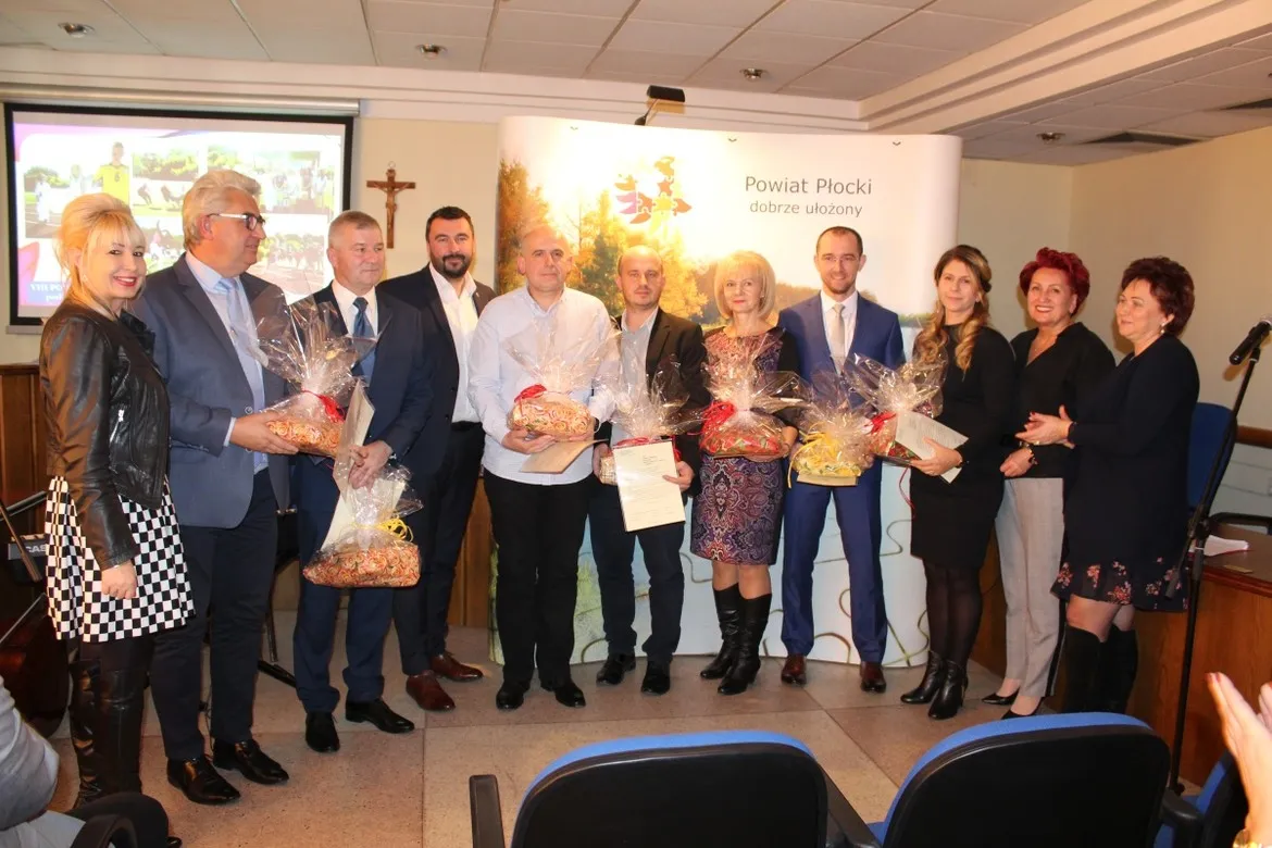 Grupa osób w eleganckich strojach stoi na sali podczas podsumowania współzawodnictwa sportowego szkół w powiecie płockim, trzymając kosze z upominkami i dokumenty.