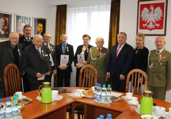 Grupa kombatantów i urzędników w formalnych strojach (część w mundurach) stoi przy okrągłym stole podczas ceremonii nadania Odznak Kombatanckich w powiecie płockim, z widocznymi medalami i dyplomami.