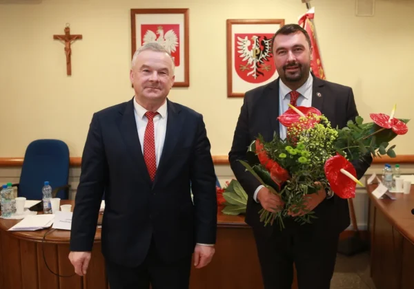 Dwóch mężczyzn w garniturach stoi w sali sesyjnej Powiatu; jeden z nich trzyma bukiet kwiatów, a w tle widoczne są polskie godła i krzyż.