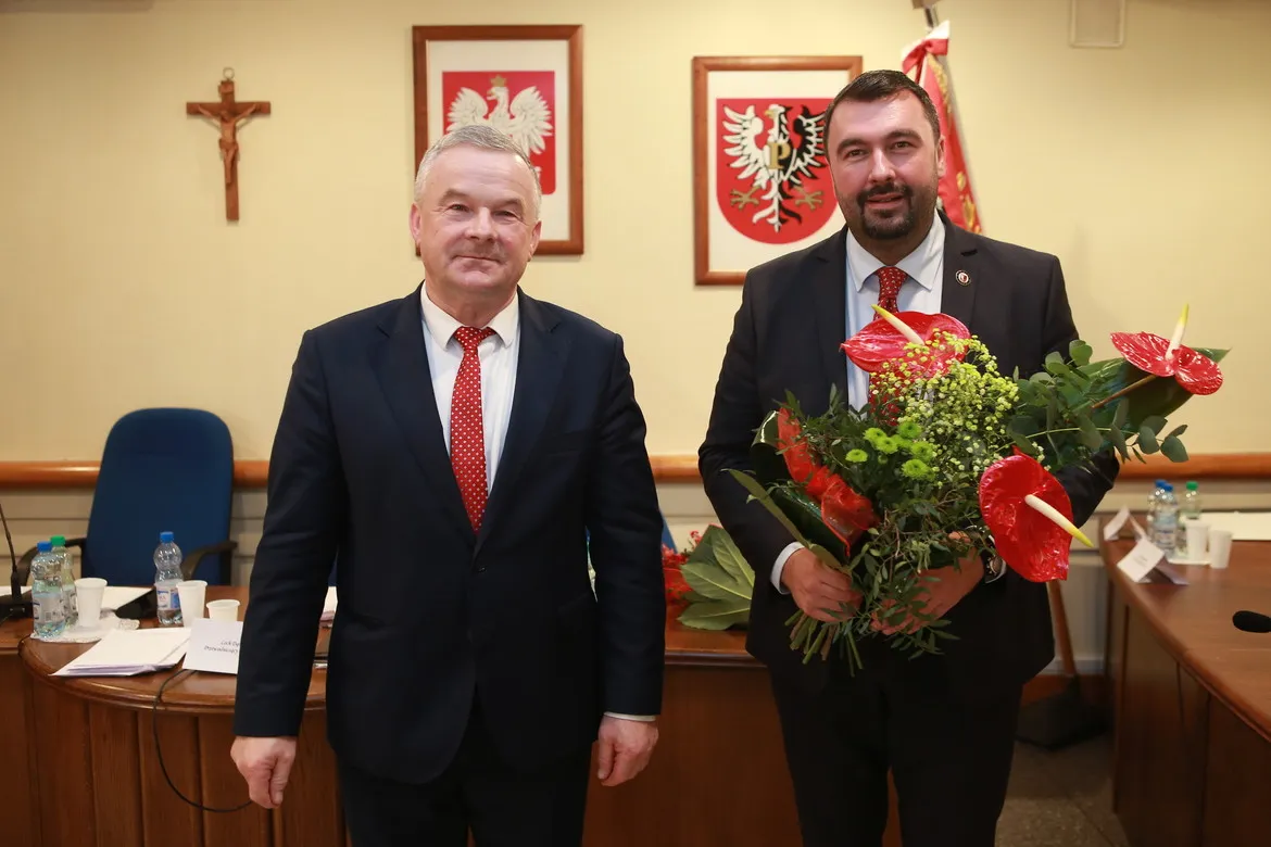 Dwóch mężczyzn w garniturach stoi w sali sesyjnej Powiatu; jeden z nich trzyma bukiet kwiatów, a w tle widoczne są polskie godła i krzyż.