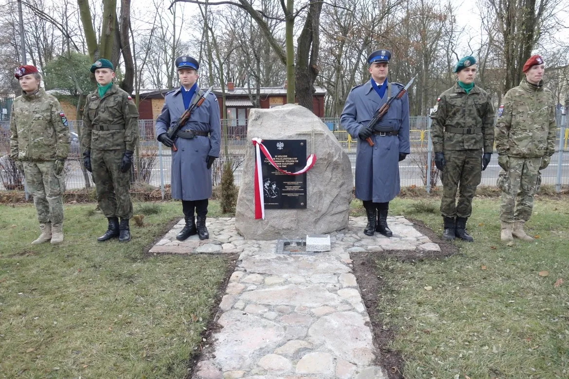 Grupa sześciu żołnierzy w różnych mundurach stoi po bokach kamiennego pomnika z tablicą, oplecionego biało‑czerwoną wstęgą, upamiętniającego płockich policjantów pomordowanych i poległych w obronie Ojczyzny.