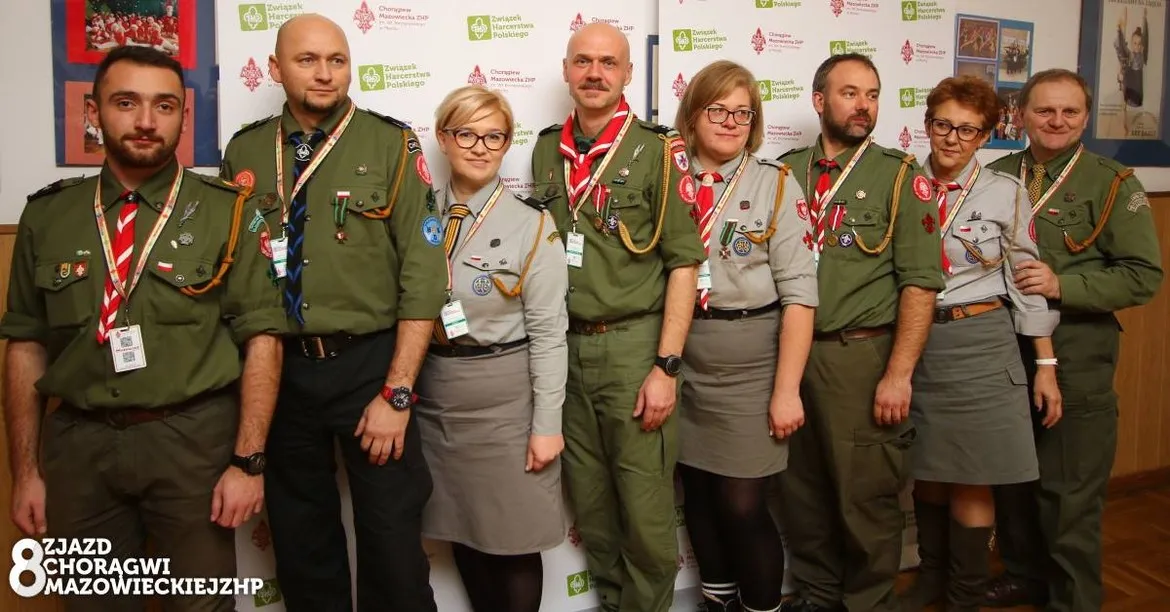 Grupa harcerzy w mundurach ZHP stoi do zdjęcia podczas Zjazdu Chorągwi Mazowieckiej ZHP, z identyfikatorami i naszywkami, w tle banery z logo ZHP.