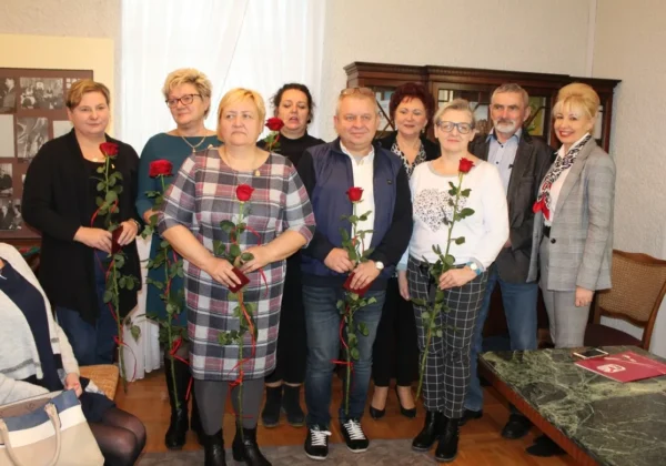 Grupa dorosłych osób stoi w wnętrzu, trzyma czerwone róże i uśmiecha się, podczas świątecznego spotkania ZNP o reformie oświaty.