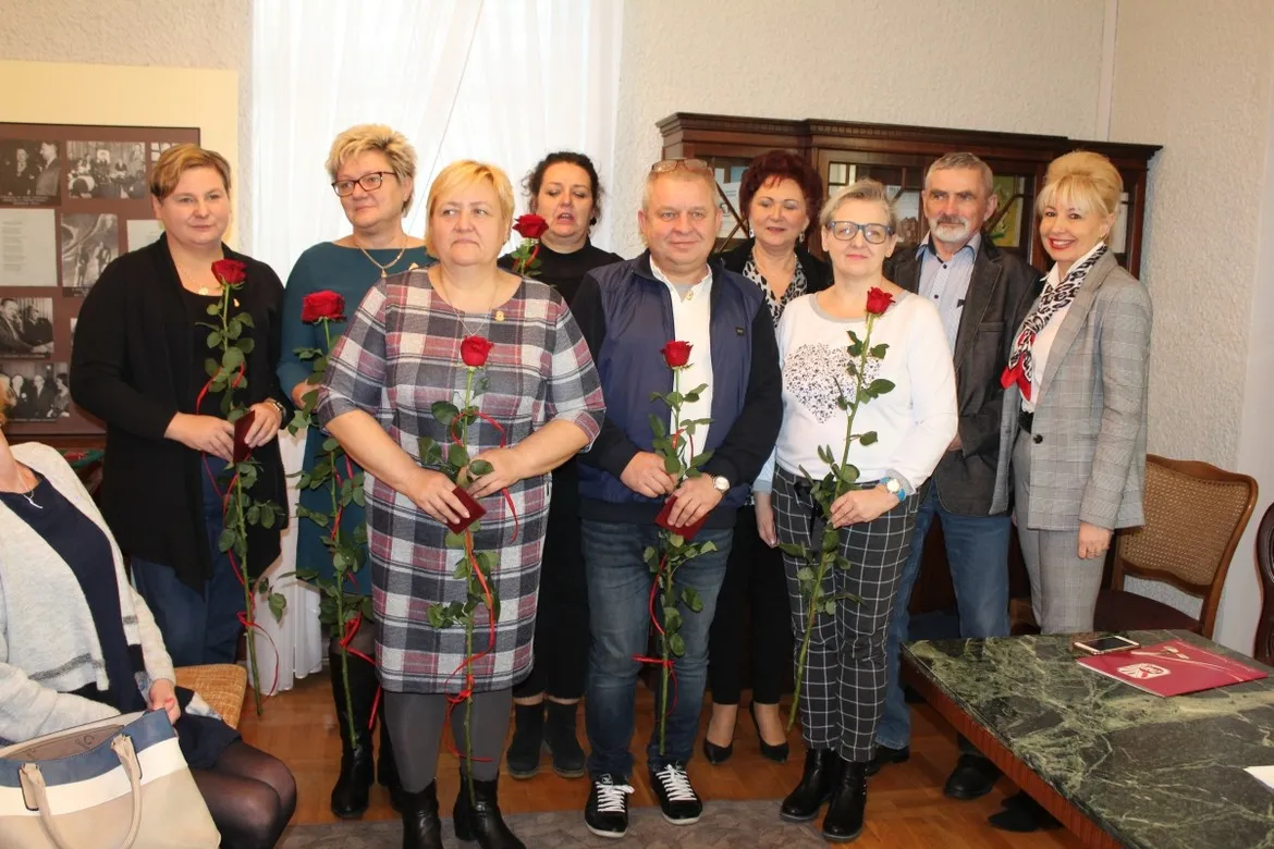 Grupa dorosłych osób stoi w wnętrzu, trzyma czerwone róże i uśmiecha się, podczas świątecznego spotkania ZNP o reformie oświaty.