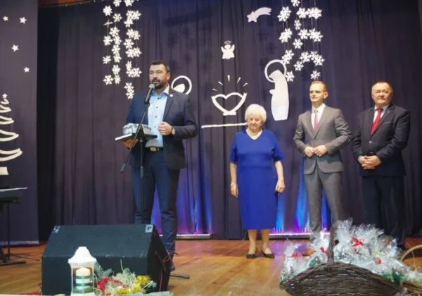 Mężczyzna w garniturze przemawia do mikrofonu na scenie podczas wieczoru wigilijnego w Radzanowie, w towarzystwie trzech osób, z zimowymi dekoracjami i koszami z prezentami na pierwszym planie.