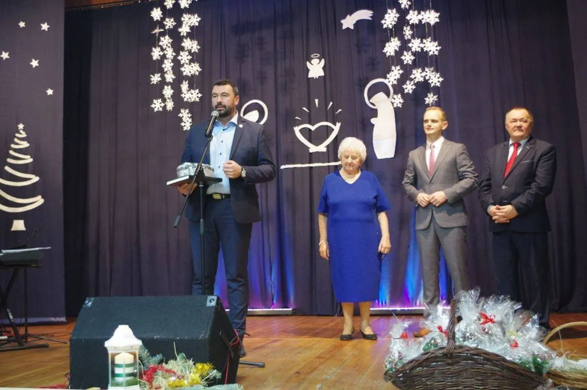 Mężczyzna w garniturze przemawia do mikrofonu na scenie podczas wieczoru wigilijnego w Radzanowie, w towarzystwie trzech osób, z zimowymi dekoracjami i koszami z prezentami na pierwszym planie.