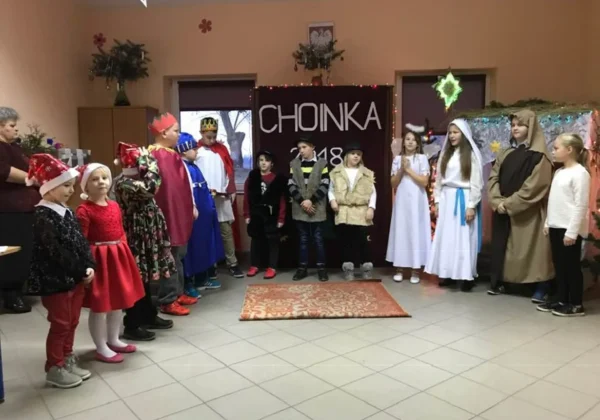 Grupa dzieci w kolorowych przebraniach (anioły, królowie, pasterze) stoi w półkolu podczas Wigilii w Świetlicy Środowiskowej w Grodkowie, przed dekorowaną sceną z napisem CHOINKA 2018.