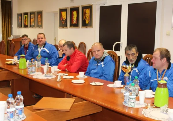 Grupa mężczyzn w niebieskich kurtkach i jeden w czerwonej bluzie siedzi przy długim stole w sali Starostwa Powiatowego w Płocku podczas wyjątkowego święta, z napojami i przekąskami na stole.