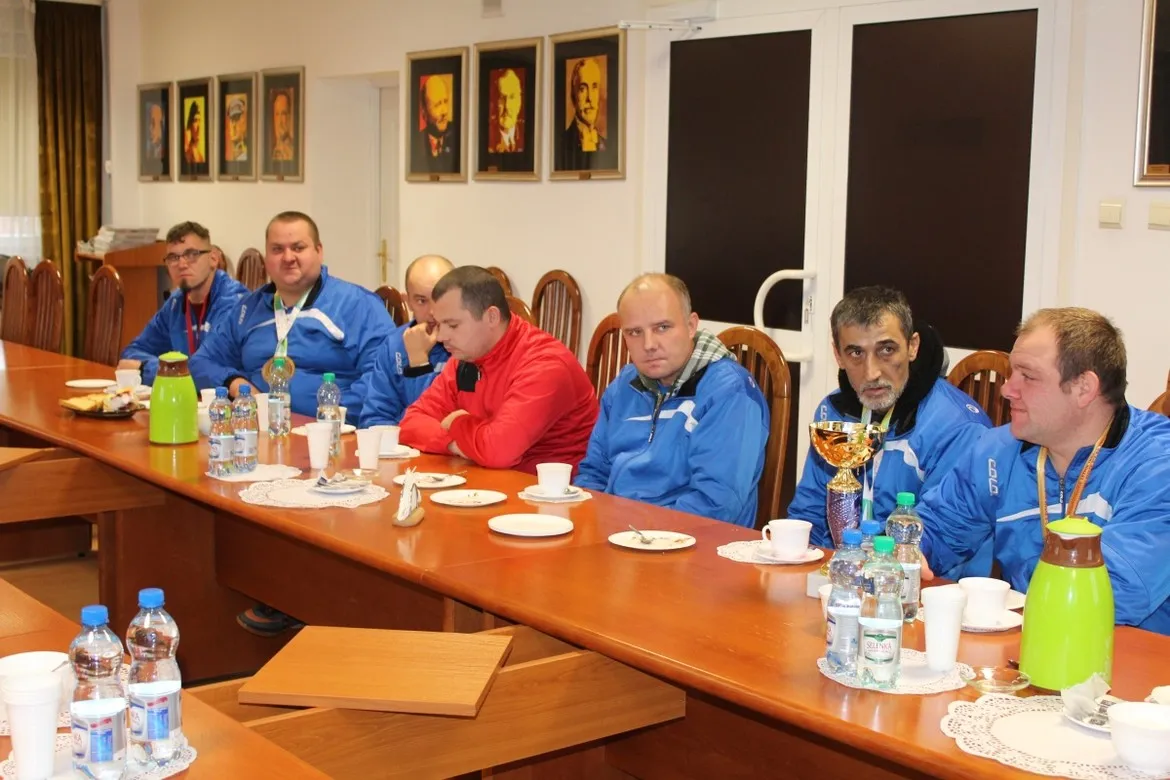 Grupa mężczyzn w niebieskich kurtkach i jeden w czerwonej bluzie siedzi przy długim stole w sali Starostwa Powiatowego w Płocku podczas wyjątkowego święta, z napojami i przekąskami na stole.