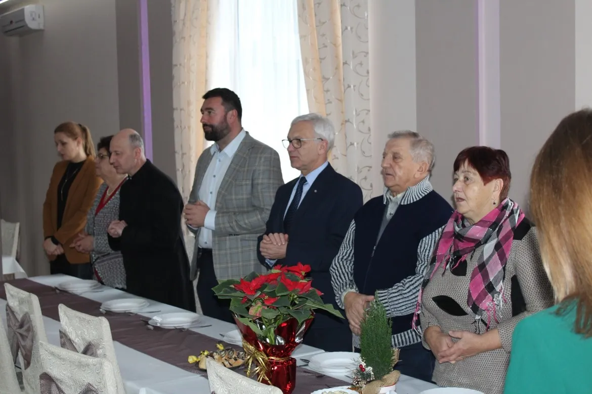 Grupa seniorów stoi przy długim stole w sali bankietowej podczas świątecznego spotkania Gminy Mała Wieś.