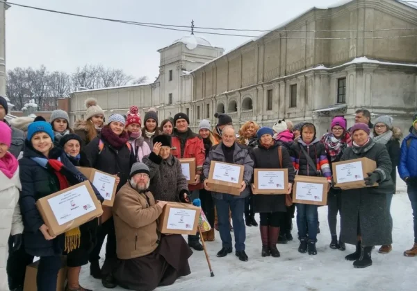 Grupa osób ubranych zimowo stoi na zaśnieżonym placu przed zabytkowym budynkiem, trzymając kartonowe pudełka z darami w ramach akcji charytatywnej „Paka Dla Rodaka”.