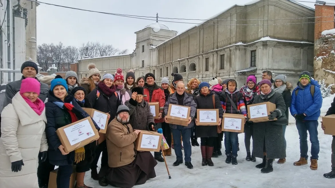 Grupa osób ubranych zimowo stoi na zaśnieżonym placu przed zabytkowym budynkiem, trzymając kartonowe pudełka z darami w ramach akcji charytatywnej „Paka Dla Rodaka”.