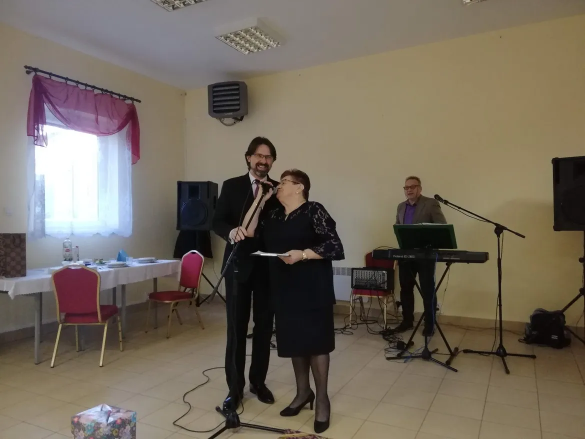 Para przy mikrofonach na scenie podczas otwarcia Klubu Senior+ w gminie Drobin, z muzykiem grającym na keyboardzie w tle.