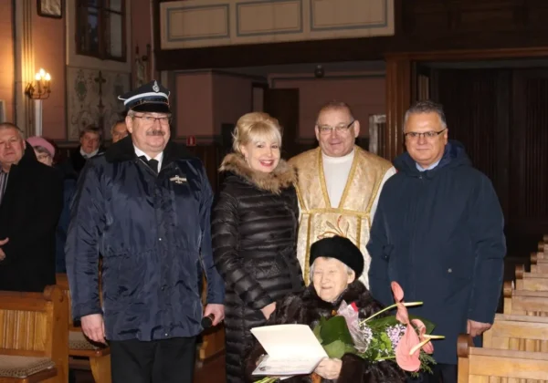 Starsza pani w futerku siedzi z bukietem i dyplomem, otoczona rodziną i duchownym podczas jubileuszu 90-lecia urodzin w kościelnym wnętrzu.