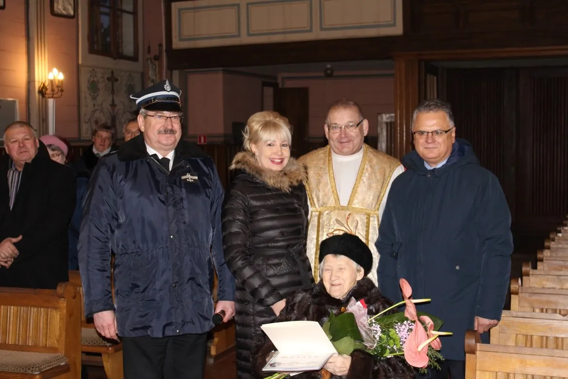 Starsza pani w futerku siedzi z bukietem i dyplomem, otoczona rodziną i duchownym podczas jubileuszu 90-lecia urodzin w kościelnym wnętrzu.