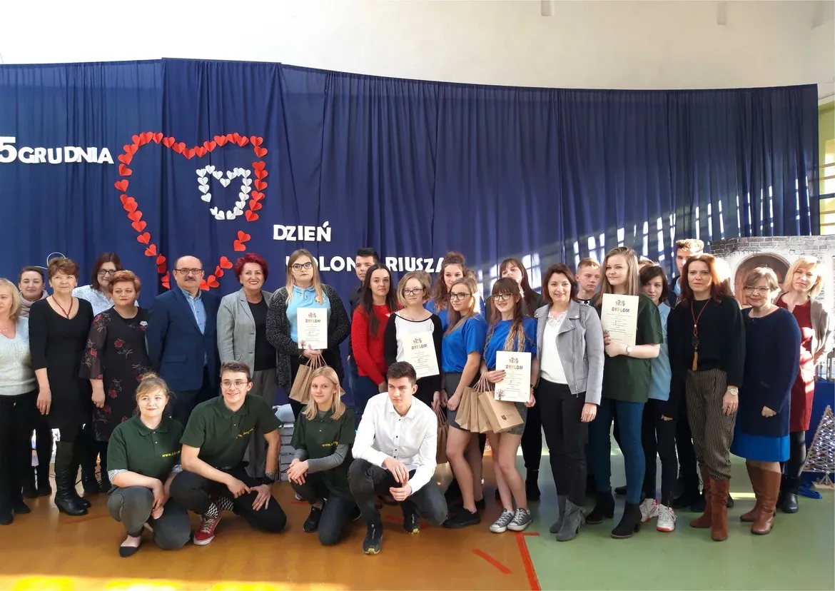 Grupa uczestników III Spotkania Integracyjnego Szkolnych Klubów Wolontariusza stoi razem na scenie przed niebieską kurtyną z napisem Dzień Wolontariuszy, część z nich trzyma dyplomy.