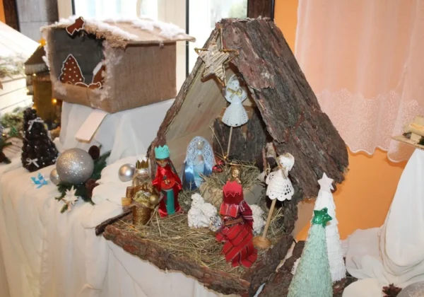 Rustykalna szopka bożonarodzeniowa z chatką wykonaną z kory i słomy, ozdobiona figurkami aniołów i królów oraz drewnianymi choineczkami, ustawiona na białej tkaninie w DPS w Koszelewie.