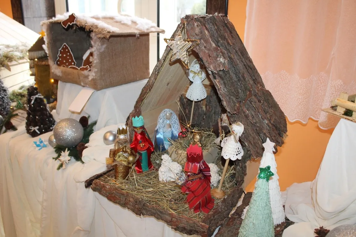 Rustykalna szopka bożonarodzeniowa z chatką wykonaną z kory i słomy, ozdobiona figurkami aniołów i królów oraz drewnianymi choineczkami, ustawiona na białej tkaninie w DPS w Koszelewie.