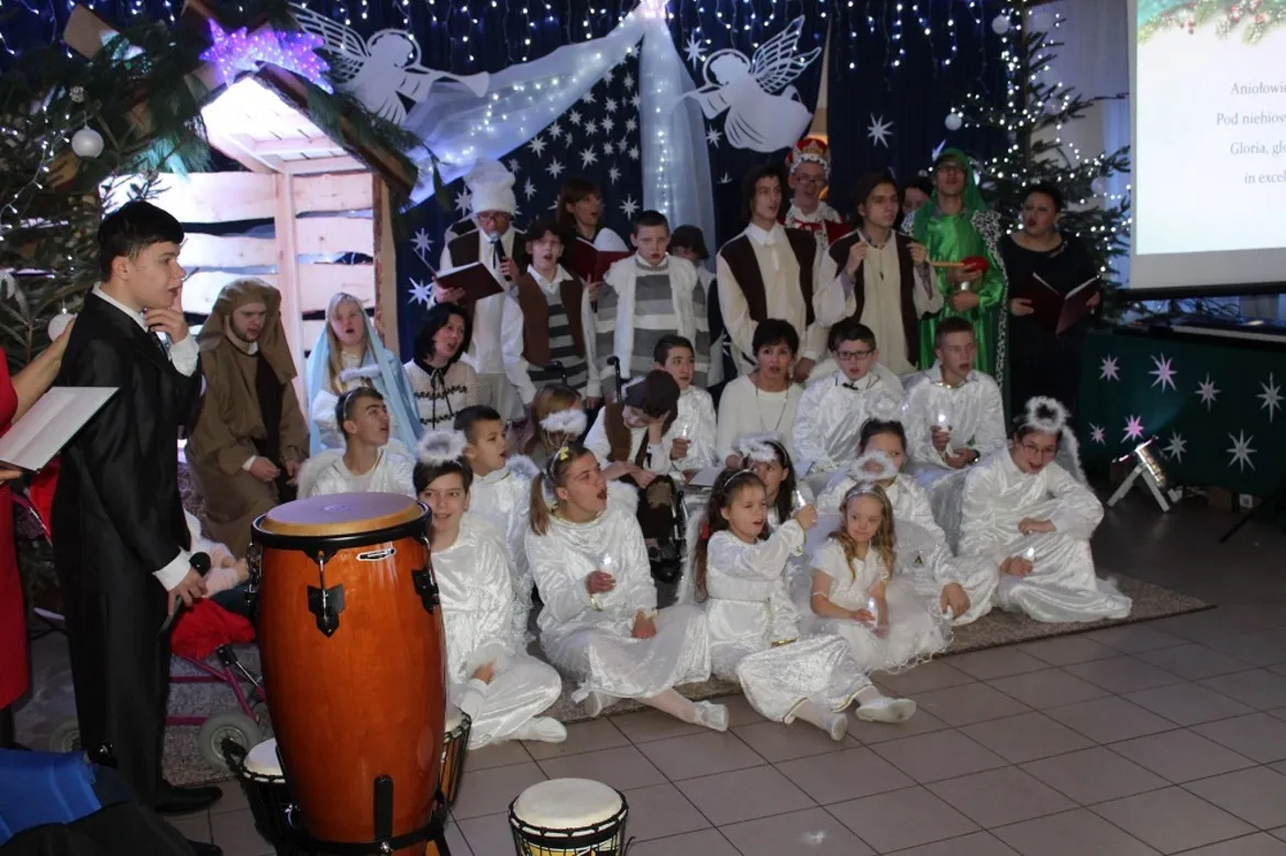 Grupa uczniów Zespołu Szkół Specjalnych w Goślicach ubranych na biało podczas opłatkowego spotkania, ustawiona w sali z bożonarodzeniowymi dekoracjami i perkusyjnymi instrumentami na pierwszym planie.