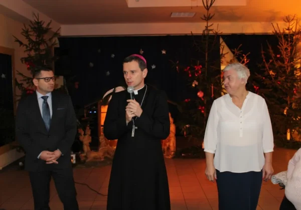 Trzy osoby stoją na sali podczas wieczerzy wigilijnej w Goślicach: ksiądz z mikrofonem w czarnej sutannie pośrodku, mężczyzna w garniturze po lewej i starsza kobieta w białej bluzce po prawej, w tle bożonarodzeniowe dekoracje i choinki.