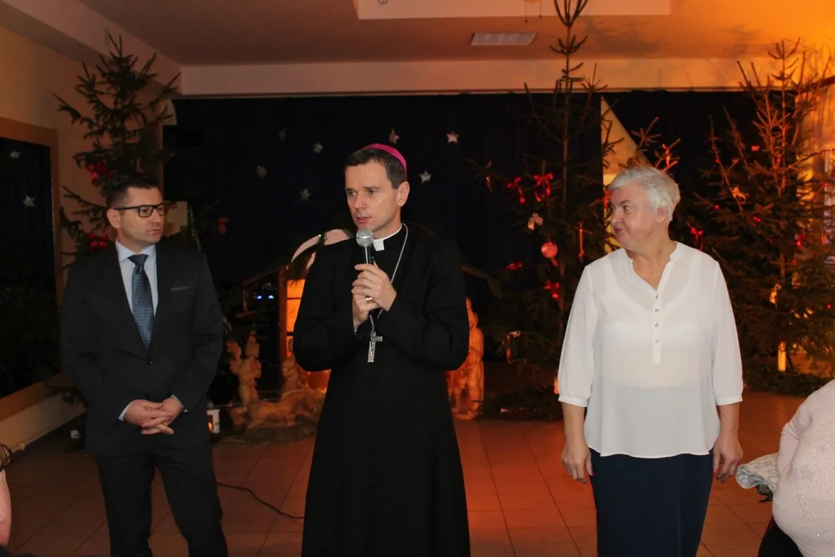 Trzy osoby stoją na sali podczas wieczerzy wigilijnej w Goślicach: ksiądz z mikrofonem w czarnej sutannie pośrodku, mężczyzna w garniturze po lewej i starsza kobieta w białej bluzce po prawej, w tle bożonarodzeniowe dekoracje i choinki.