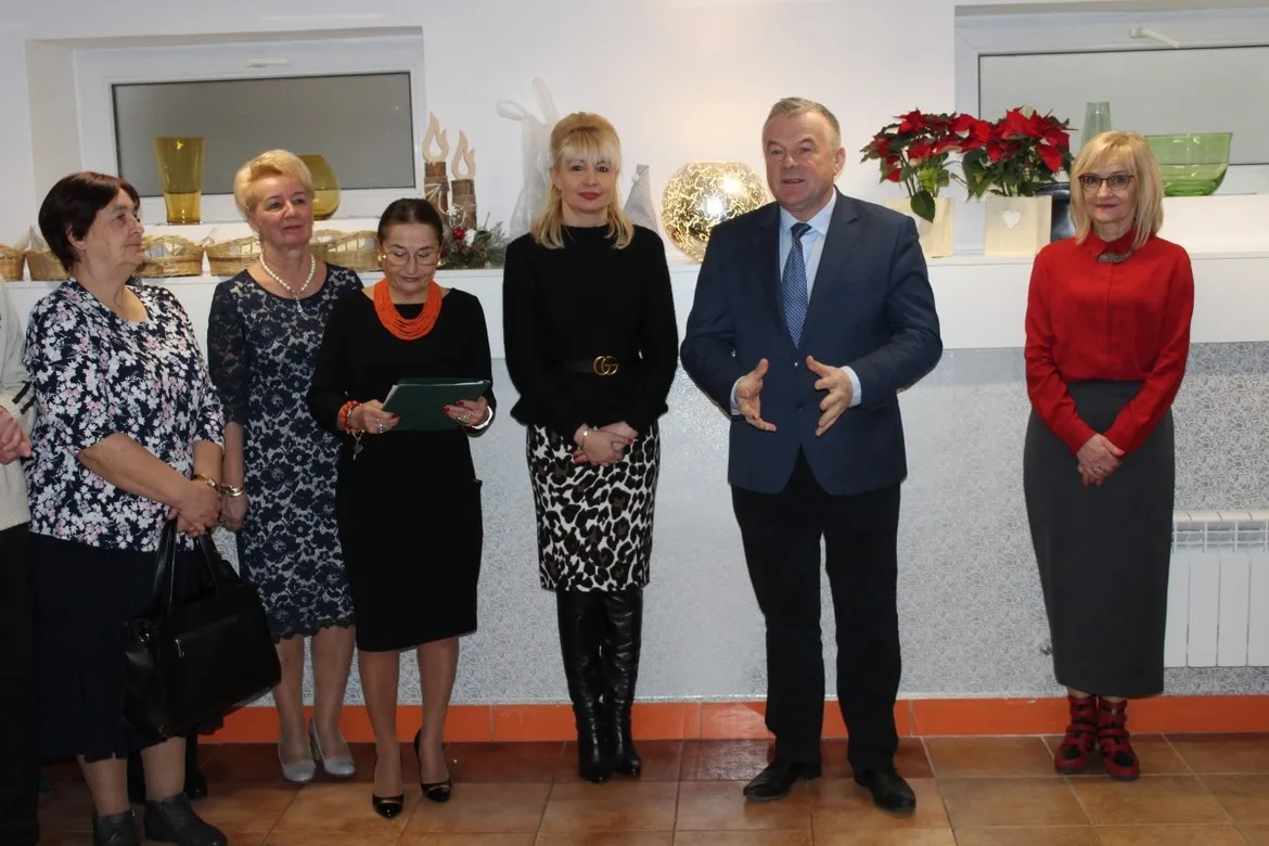 Grupa sześciu osób w eleganckich strojach stoi w holu Domu Pomocy Społecznej Nad Jarem w Nowym Miszewie podczas spotkania opłatkowego.