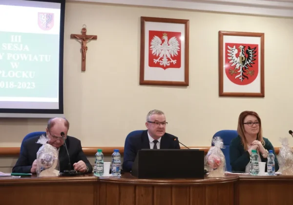 Trzech urzędników siedzi przy stole w sali obrad podczas III sesji Rady Powiatu w Płocku, gdy uchwalono przyszłoroczny budżet; w tle widoczny projektor z napisem sesji oraz herby powiatu i krzyż.