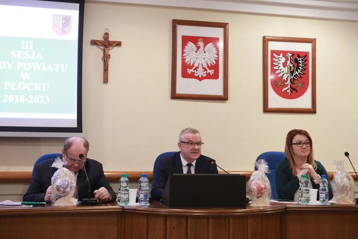 Trzech urzędników siedzi przy stole w sali obrad podczas III sesji Rady Powiatu w Płocku, gdy uchwalono przyszłoroczny budżet; w tle widoczny projektor z napisem sesji oraz herby powiatu i krzyż.