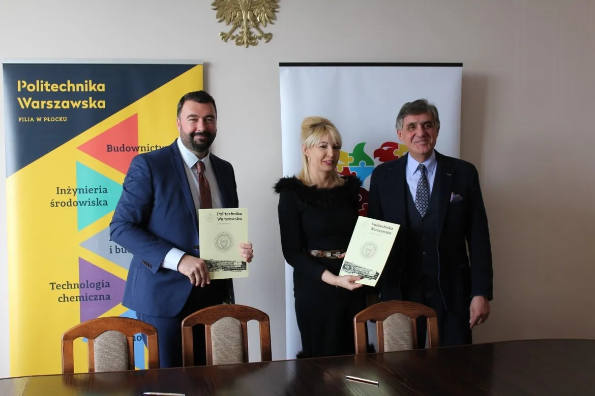 Trzy osoby w garniturach stoją przy stole, trzymają dokumenty z logo Politechniki Warszawskiej, na tle bannerów uczelni.