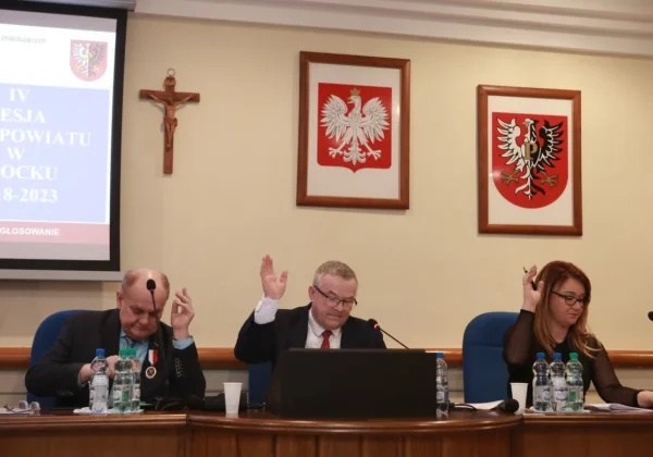 Trzech samorządowców siedzi za stołem podczas IV sesji Rady Powiatu, wszyscy podnoszą rękę w trakcie głosowania, z projektorem i herbami na ścianie w tle.