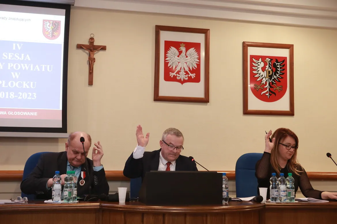 Trzech samorządowców siedzi za stołem podczas IV sesji Rady Powiatu, wszyscy podnoszą rękę w trakcie głosowania, z projektorem i herbami na ścianie w tle.