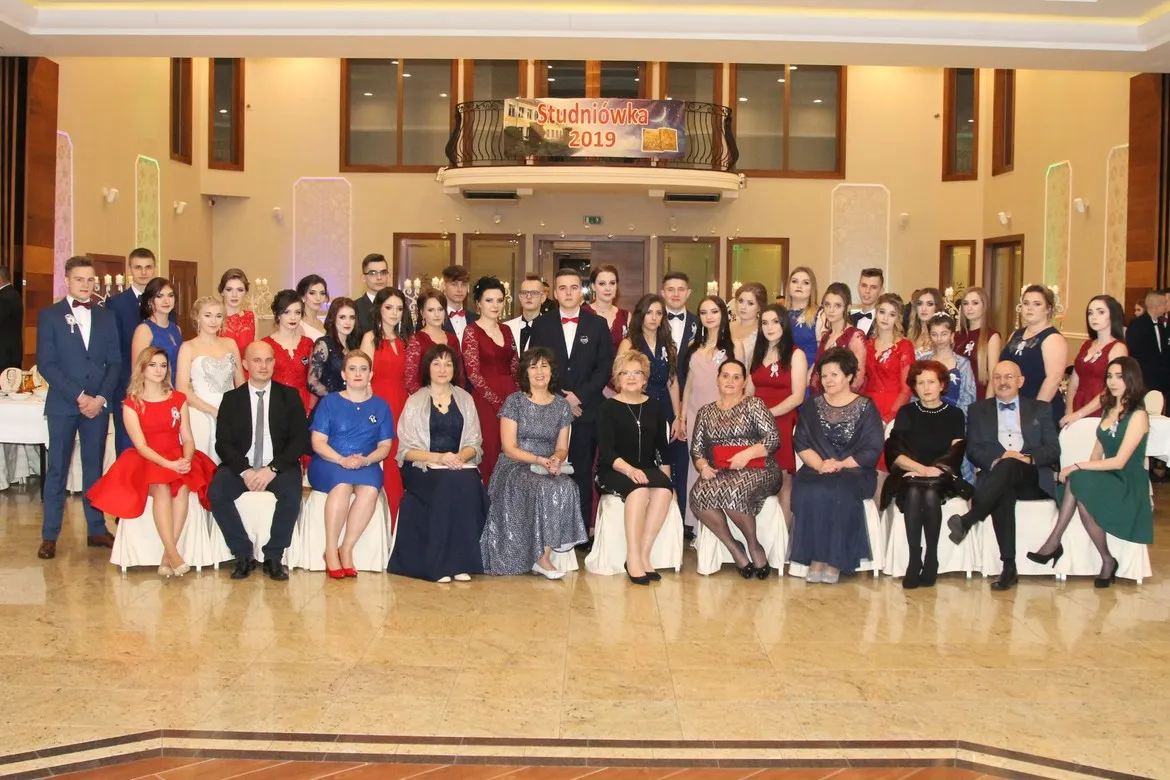 Grupa uczniów i nauczycieli Zespołu Szkół im. St. Staszica w Gąbinie podczas Studniówki 2019, ustawiona w sali balowej przed banerem „Studniówka 2019”.