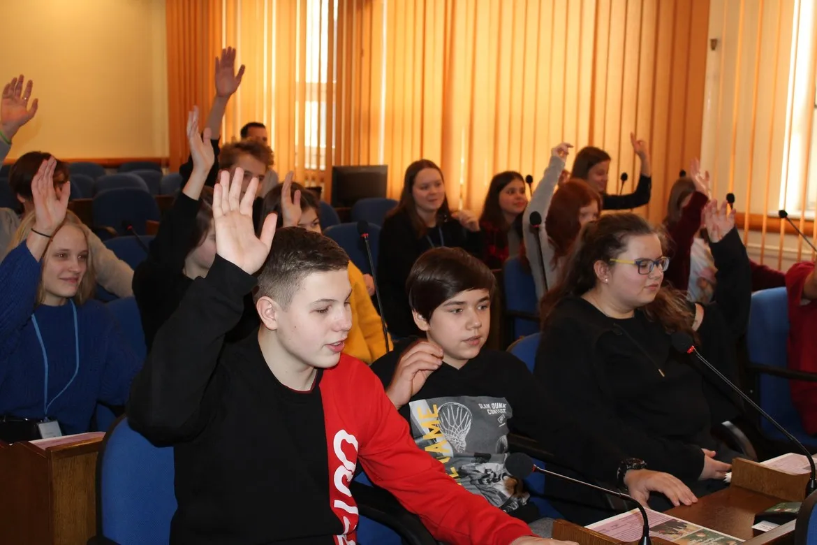 Uczniowie Ogólnokształcącej Szkoły Muzycznej podczas wizyty w Starostwie siedzą w sali konferencyjnej; część z nich podnosi ręce, a przed nimi znajdują się mikrofony.