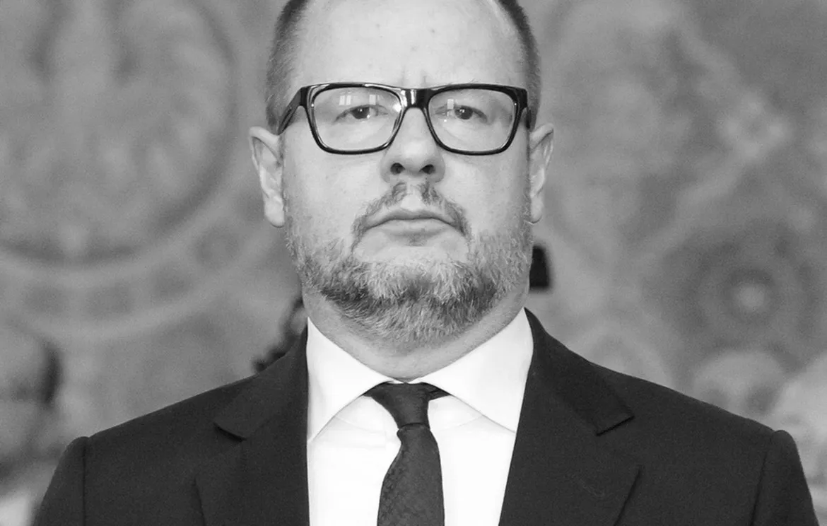 Czarno-białe zdjęcie portretowe mężczyzny w garniturze i okularach o poważnym wyrazie twarzy na rozmytym tle.