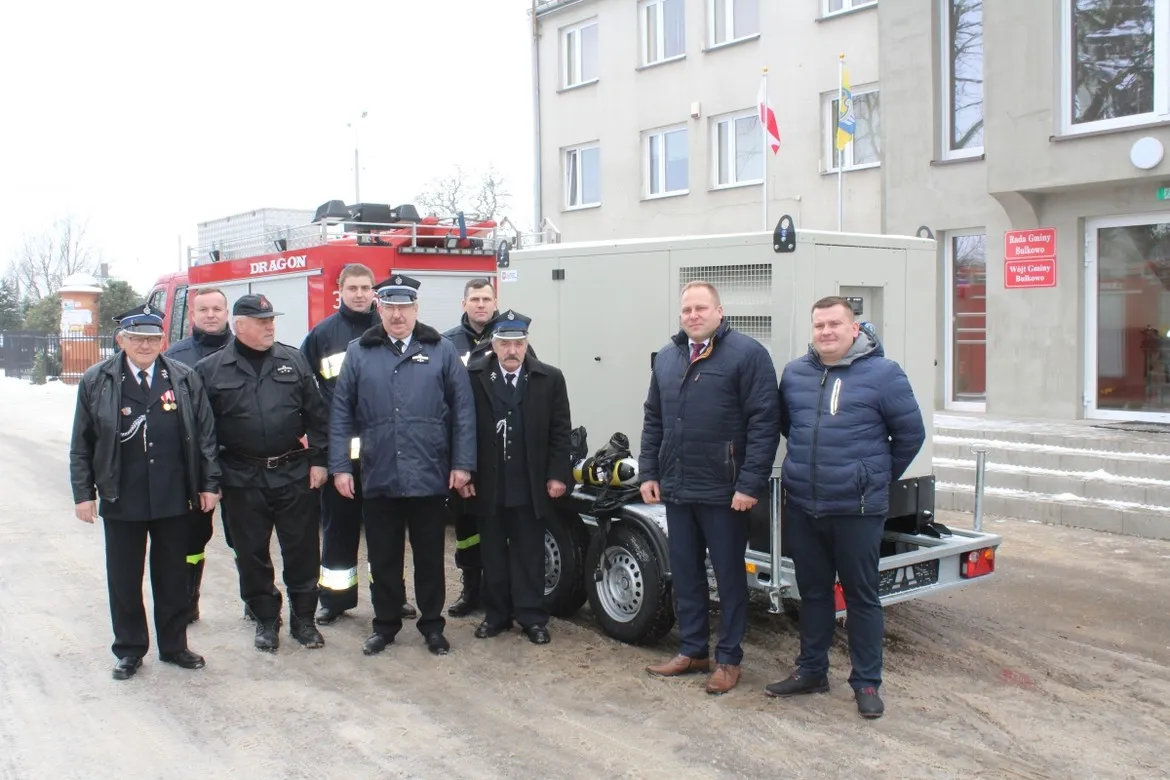 Grupa osób, w tym strażacy i urzędnicy, stoi obok przyczepy z wyposażeniem sprzętu specjalistycznego podczas przekazania go jednostkom z Gminy Bulkowo.