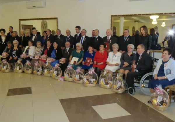 Grupowe zdjęcie z uroczystości odznaczeń dla małżeństw z gminy Słubice: osoby w formalnych strojach stoją i siedzą, część z medalami i dyplomami, a przed nimi ułożone kosze upominkowe.