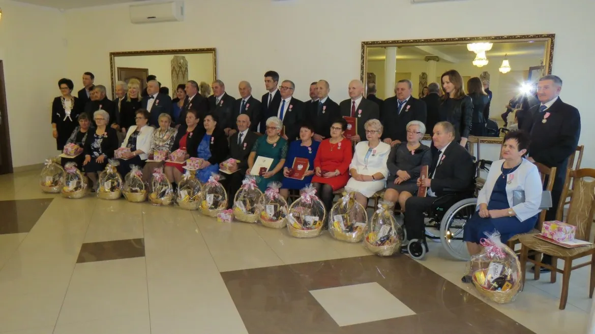 Grupowe zdjęcie z uroczystości odznaczeń dla małżeństw z gminy Słubice: osoby w formalnych strojach stoją i siedzą, część z medalami i dyplomami, a przed nimi ułożone kosze upominkowe.