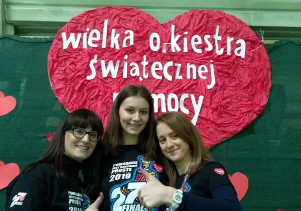 Trzy uśmiechnięte kobiety stojące przed dużym czerwonym sercem z napisem „Wielka Orkiestra Świątecznej Pomocy” podczas WOŚP.