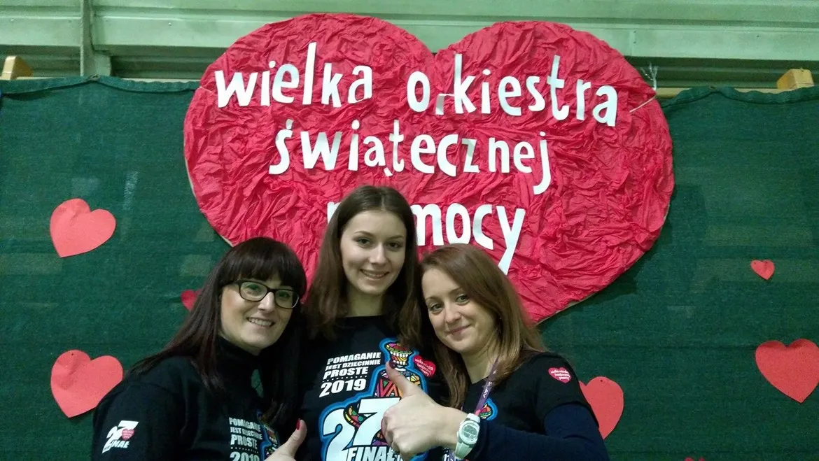 Trzy uśmiechnięte kobiety stojące przed dużym czerwonym sercem z napisem „Wielka Orkiestra Świątecznej Pomocy” podczas WOŚP.