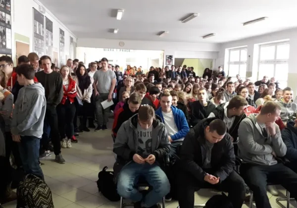 Duża grupa uczniów zgromadzona w szkolnej auli, część siedzi na krzesłach, inni stoją w tle.