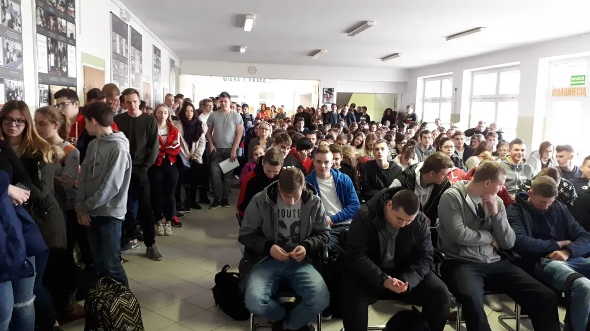 Duża grupa uczniów zgromadzona w szkolnej auli, część siedzi na krzesłach, inni stoją w tle.