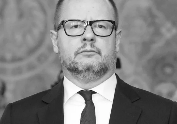 Portret mężczyzny w czarnym garniturze i krawacie, noszącego okulary, na czarno-białym tle.